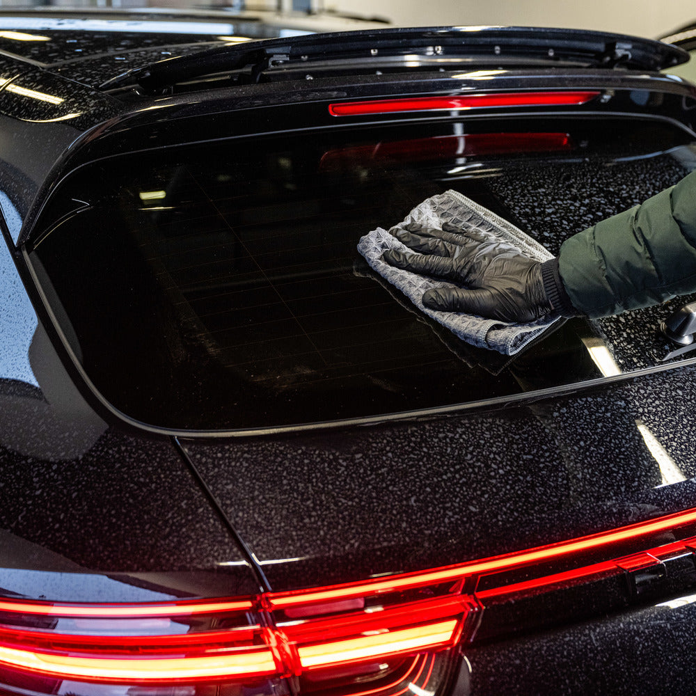 Using the Interior Sample Package, a gloved person cleans the wet rear window of a black car with a microfiber cloth while its red taillights are illuminated.