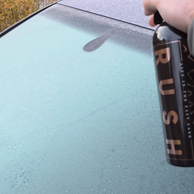 A hand sprays De-Icer onto a car's glass roof, showing water beading on the smooth, clean surface.