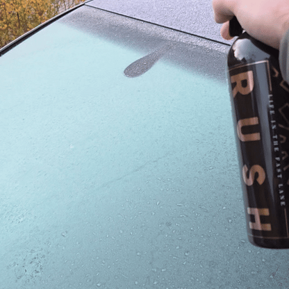A hand sprays De-Icer onto a car's glass roof, showing water beading on the smooth, clean surface.