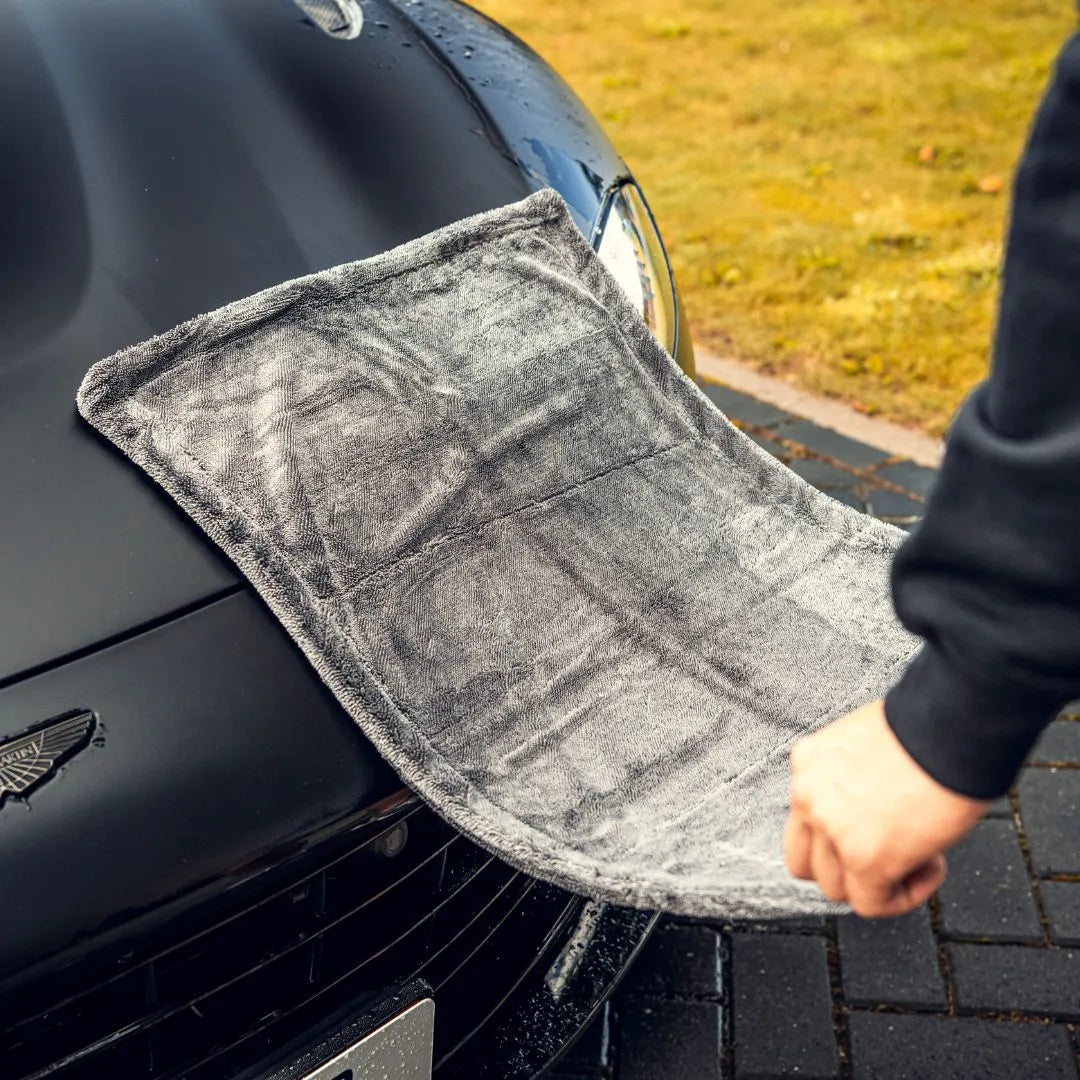 A hand wipes the hood of a black car with the Master Drying Towel 1200 GSM. Parked near grass, the car’s spotless finish shows this high-GSM doek’s impressive drying power for auto care.