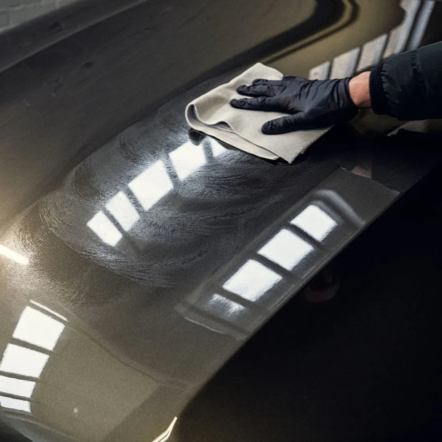 Wearing a black glove, a person uses a light cloth and 1+1 GRATIS Spray Wax to polish the hood of a dark car, enhancing lak bescherming and reflecting bright overhead lights in a garage or workshop.