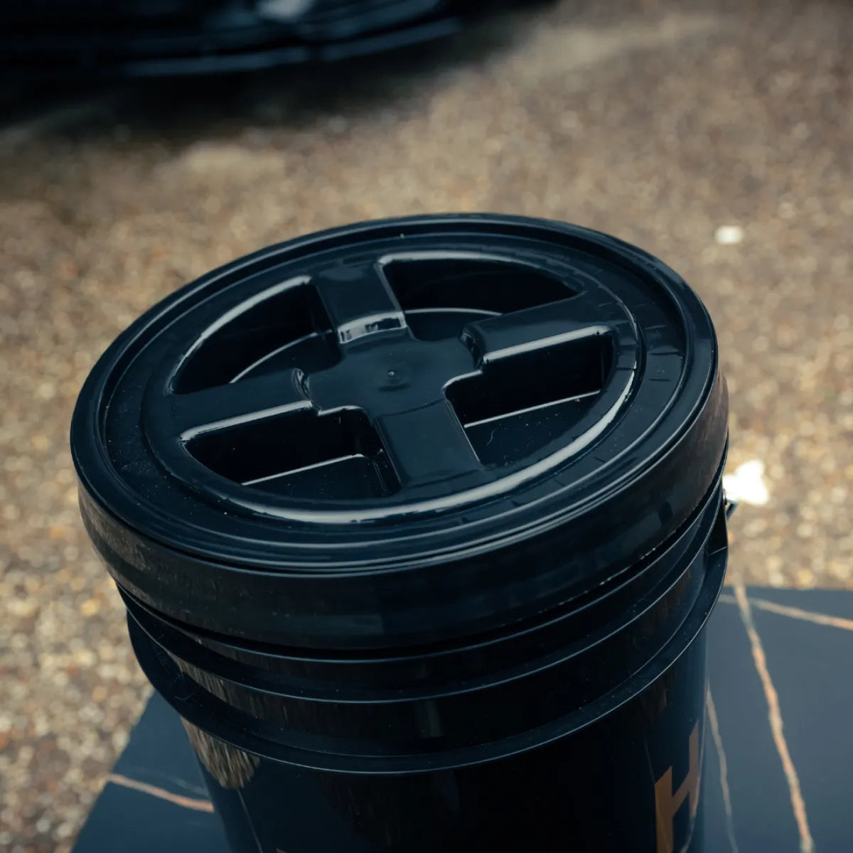 A close-up of a black Detailing Bucket 20L with a cross-shaped lid, placed outdoors on gravel and pavement. Ideal for the two-bucket method or adding a Grit Guard for optimal car care.