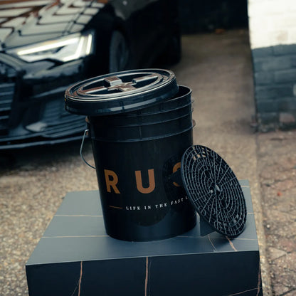 A Bonus Bucket Kit labeled “RUSH” with “LIFE IN THE FAST LANE” sits on a gray box outdoors. Its black closed lid and a black grit guard are visible, with a black car in the background.