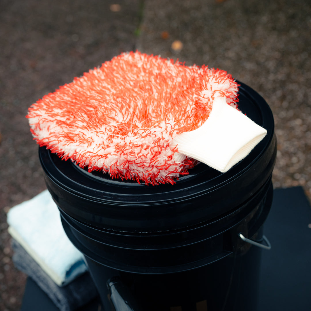 An orange and white chenille wash mitt rests on a black Bonus Bucket Kit, with two folded towels visible in the background on the left.