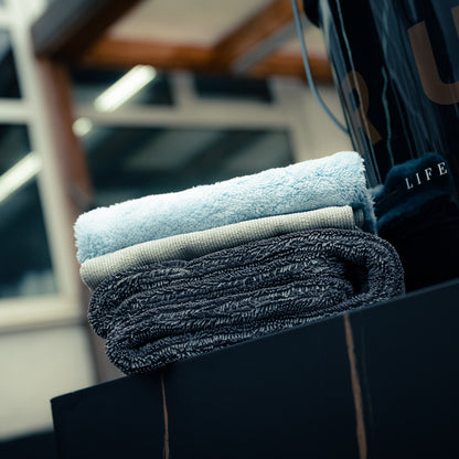 Two neatly folded towels, one light blue and one dark gray, are stacked on a black surface indoors next to a Bonus Bucket Kit, with large windows and a blurred background in view.
