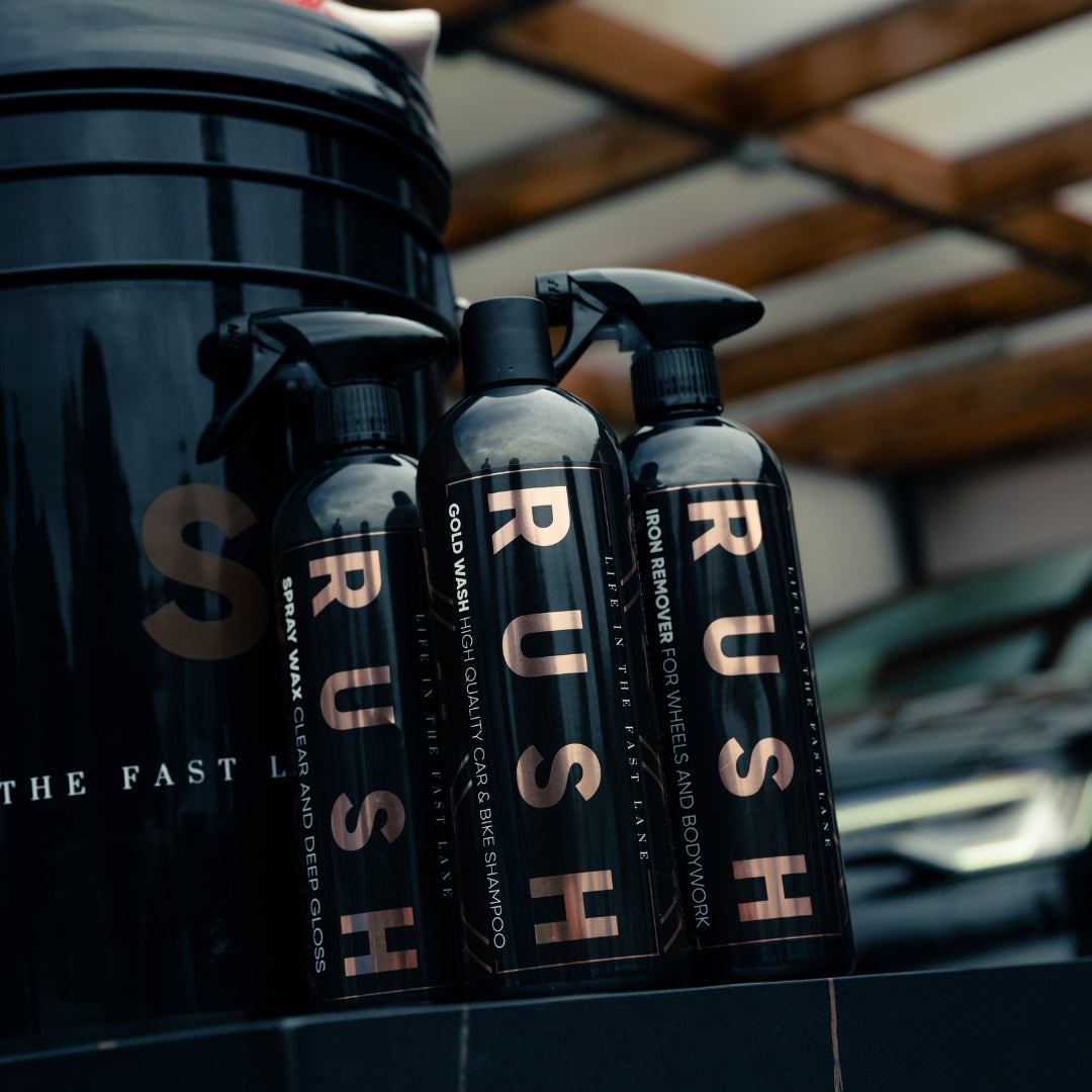 Three black spray bottles labeled RUSH Iron Remover are arranged side by side with a large black container, set against a background featuring a car and wooden roof structure—showcasing the Bonus Bucket Kit.