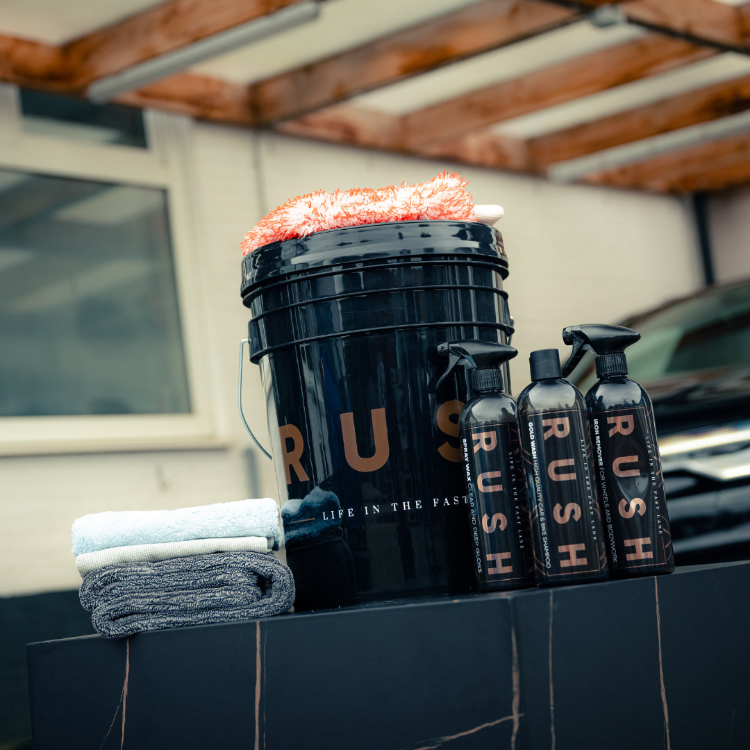 The Bonus Bucket Kit, labeled RUSH, contains an orange wash mitt, two folded towels, and three bottles—RUSH Gold Wash and RUSH Iron Remover included—arranged outdoors next to a car and building.