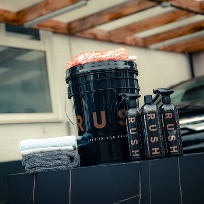 The Bonus Bucket Kit, labeled RUSH, contains an orange wash mitt, two folded towels, and three bottles—RUSH Gold Wash and RUSH Iron Remover included—arranged outdoors next to a car and building.