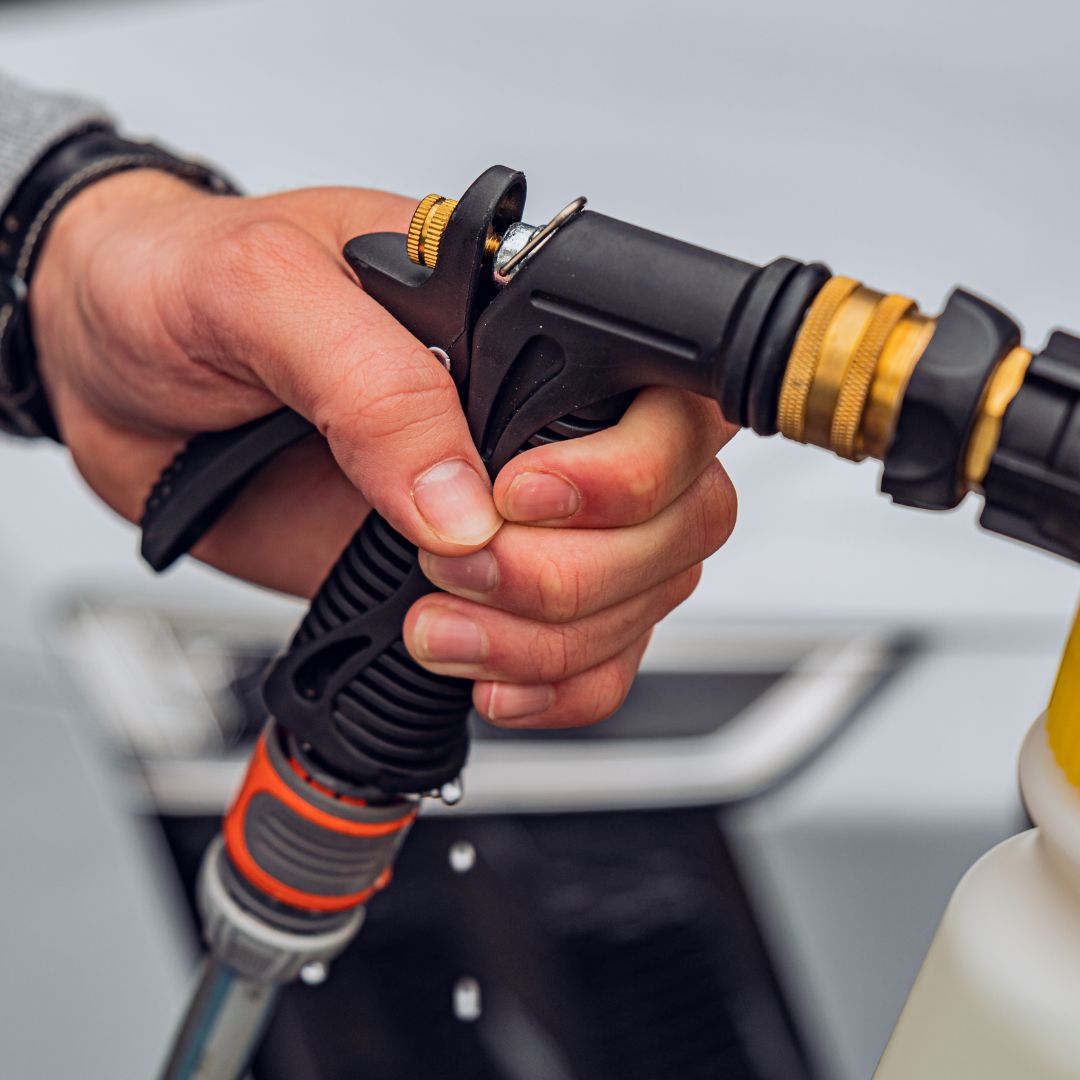 A close-up of a person’s hand pressing the trigger on the Foam Spray Gun Package, showing its black and yellow design attached to a hose with water droplets visible near the nozzle tip.