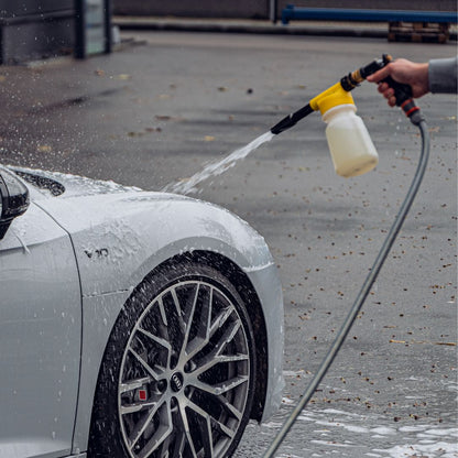 A person uses the Foam Spray Gun Package to spray water and soap onto the front side of a white car on a wet outdoor surface, with thick suds covering the wheel and part of the hood.