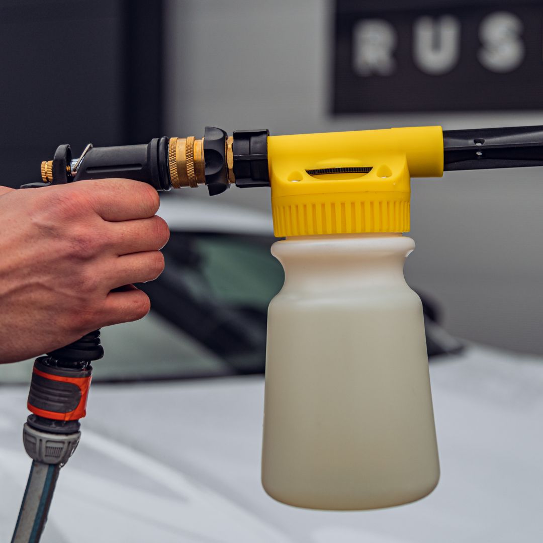 A hand holds the Foam Spray Gun Package—yellow and black nozzle with a white plastic bottle attached to a hose—shown in front of a blurred white car, ready for auto voorwas.