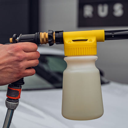 A hand holds the Foam Spray Gun Package—yellow and black nozzle with a white plastic bottle attached to a hose—shown in front of a blurred white car, ready for auto voorwas.