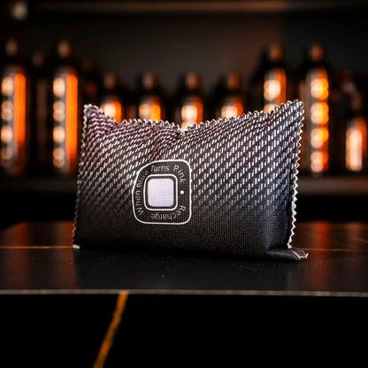 A rectangular, black textured pouch from the Winter Essentials Package, featuring a circular label, sits on a dark surface with rows of softly lit bottles in the background creating a warm atmosphere.