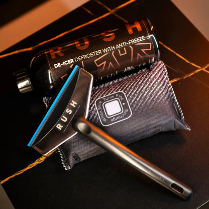 The Winter Essentials Package, featuring a black RUSH De-Icer Defroster can, a RUSH-branded black ice scraper, and a textured black pouch, is displayed on a dark marble-patterned surface.