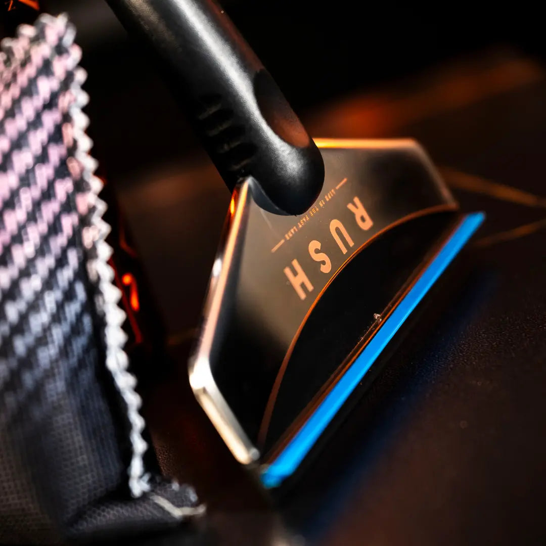 A close-up of the IJskrabber, a black and silver aluminium ice scraper with "RUSH" on it, lies beside a black-and-white checkered cloth on a dark surface—ideal for clearing ice from car windows.