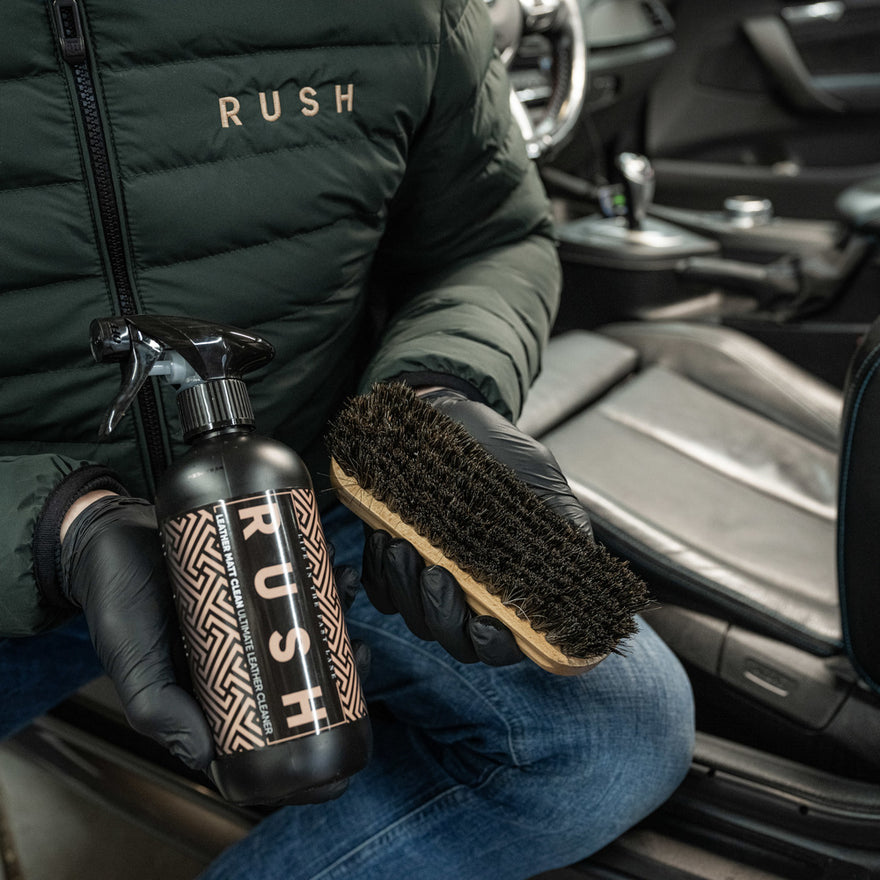 A person wearing black gloves and a green RUSH jacket holds the Leather Clean & Care Package, including cleaner and a black-bristled brush, ready to clean while sitting by a car’s leather interior.