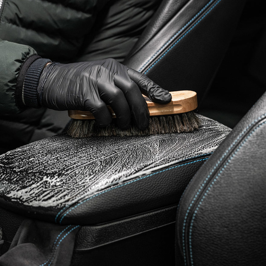 Wearing black gloves, a person uses the Leather Clean & Care Package with a wooden-handle bristle brush to clean and care for leather car seats featuring blue stitching, working up soap on the surface.