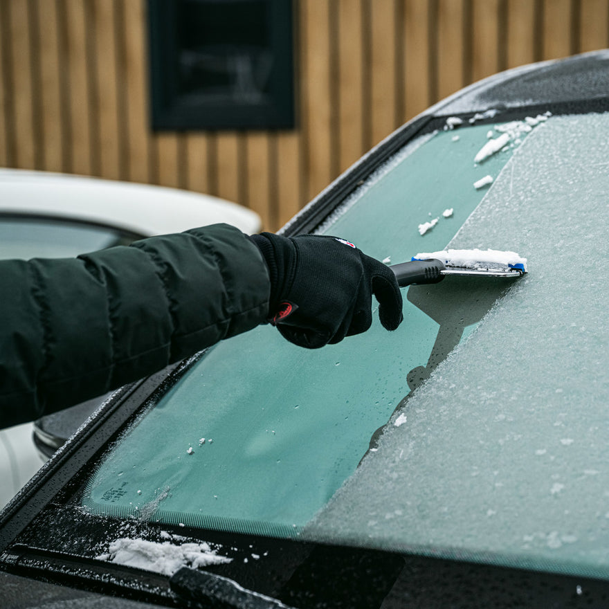 A person uses the IJskrabber to remove frost from a car windshield, making the autoruiten ijsvrij on a cold day.