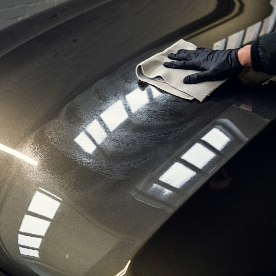 A gloved hand wipes a microfiber cloth across a dark car hood, using the 1+1 GRATIS Spray Wax Package to polish and protect the surface. Reflected light highlights swirl marks and reveals a glossy, protective wax layer.