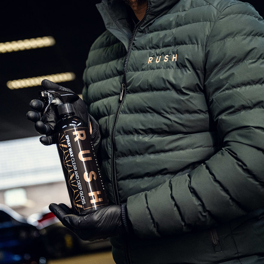 A person in a green puffer jacket and black gloves holds a black bottle from the 1+1 GRATIS Spray Wax Package indoors, with cars behind, ready to give any vehicle extra shine and a protective wax layer.