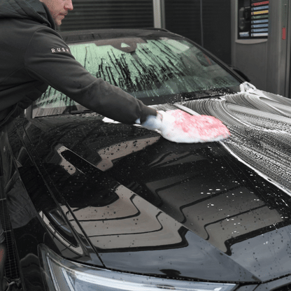 Wearing a dark jacket, a person uses a red and white sponge with autoshampoo from the Winter Protect DEAL to wash the soapy hood of an indoor, black car. Its windshield is covered in soap—ideal for winter protection.