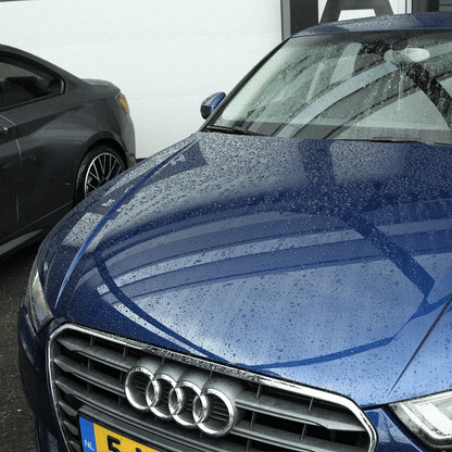 A blue Audi with rain droplets sits beside a gray car in front of a white building. The Drying Towel 600 GSM is ideal for keeping your car spotless—perfect for shining the grille and Audi logo after rain.