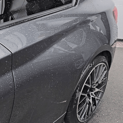 Close-up of a black car’s rear side and wheel, covered in raindrops. Treated with the Black Friday Package, the car gleams on a paved surface as reflections play across its glossy finish.