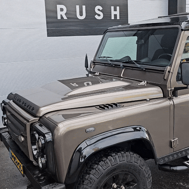 A beige Land Rover Defender with black accents is parked by a corrugated wall with a RUSH sign, gleaming after a wash using the Black Friday Package. The hood, grille, and front left side of the vehicle are clearly visible.