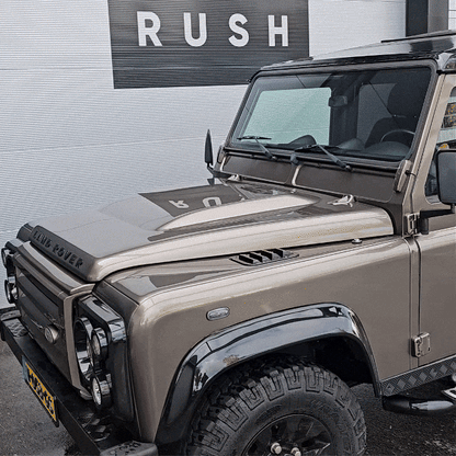 A beige Land Rover Defender with black accents is parked by a corrugated wall with a RUSH sign, gleaming after a wash using the Black Friday Package. The hood, grille, and front left side of the vehicle are clearly visible.