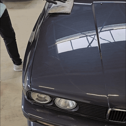 A gloved person uses a cloth to polish a dark car hood with the 1+1 GRATIS Spray Wax Package, producing auto shine and clear reflections of a building. Only their arm and part of one leg are visible in the image.