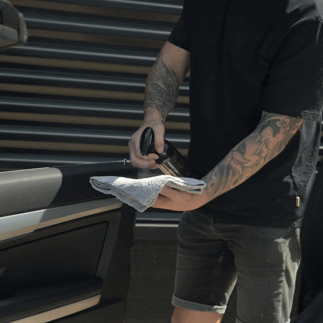 Using the Valentijnsdag Package Interior Clean, a tattooed person in casual clothing stands outside and carefully cleans the car door interior with a spray bottle and cloth.