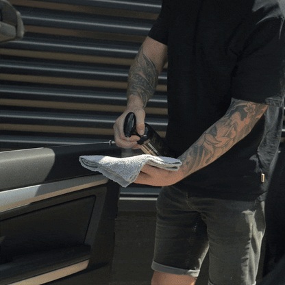 Using the Valentijnsdag Package Interior Clean, a tattooed person in casual clothing stands outside and carefully cleans the car door interior with a spray bottle and cloth.