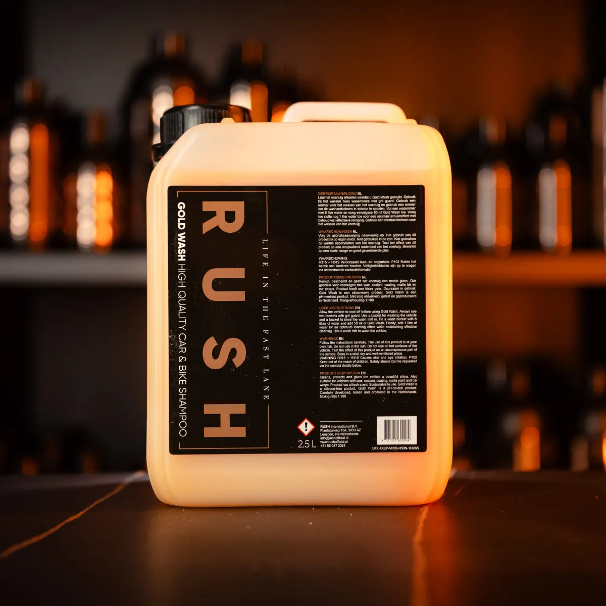 A large 2.5L Gold Wash 2,5 liter container rests on a dark surface, with shelves of similar bottles softly blurred in the background and warm lighting illuminating the scene.