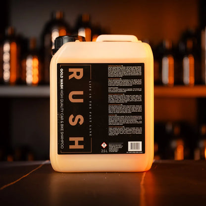 A large 2.5L Gold Wash 2,5 liter container rests on a dark surface, with shelves of similar bottles softly blurred in the background and warm lighting illuminating the scene.
