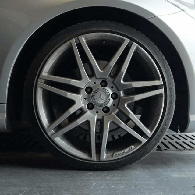 As a silver car’s wheel rim rotates indoors on a metal grate, the brake caliper and disc are revealed—ideal areas for Iron Remover to tackle tough grime before the wheel returns to its original position.