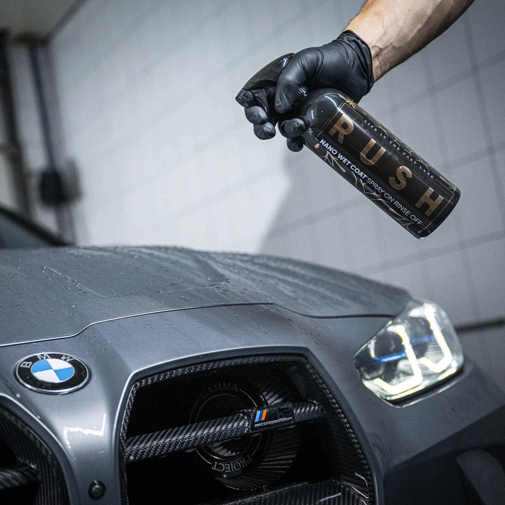A gloved hand holds a spray bottle labeled 1+1 GRATIS Nano Wet Coat near the hood of a gray BMW, preparing to apply a waterafstotende laag. The wet car is parked indoors with tiled walls in the background.