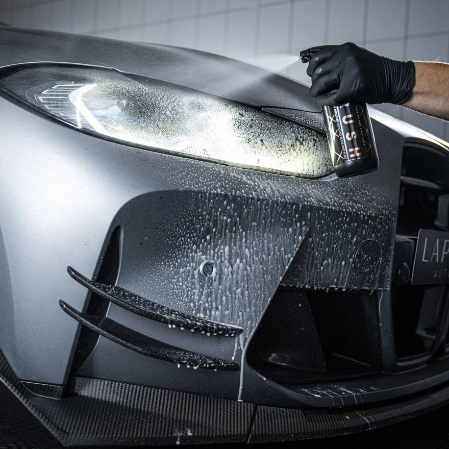 A gloved hand sprays Bug Remover onto the headlight and front bumper of a modern car, creating foam and droplets during the auto detailing process.