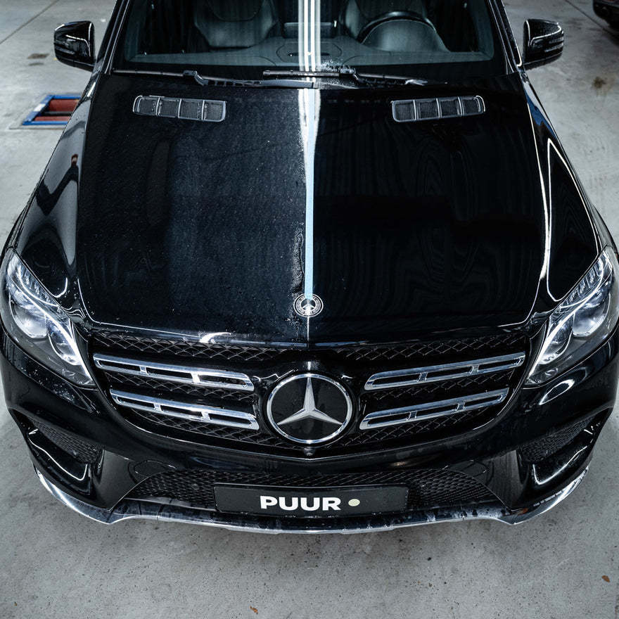 A black Mercedes-Benz in a garage shows a bold dirt line down the hood, highlighting the dramatic effect of the Ceramic Sealant Package. The PUUR license plate underscores the contrast between protected and unprotected sides.