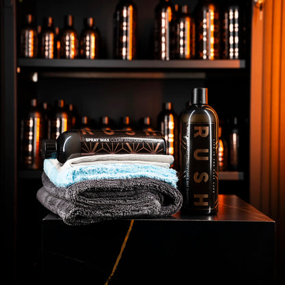 The Showroom Finish Package, featuring Spray Wax and a neatly folded microfiber droogdoek, is displayed on a black surface. Behind them, shelves lined with matching black bottles and Gold Wash create a warmly lit, glossy backdrop.