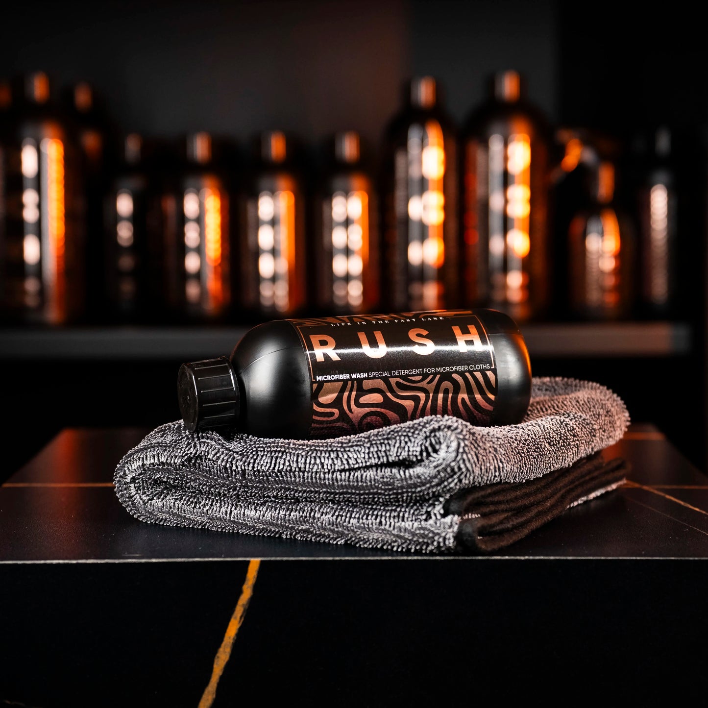 A black Perfect Drying Package bottle rests on folded gray microfiber towels, with more bottles blurred behind. The modern display and warm lighting emphasize the premium quality of this microfiber care product.