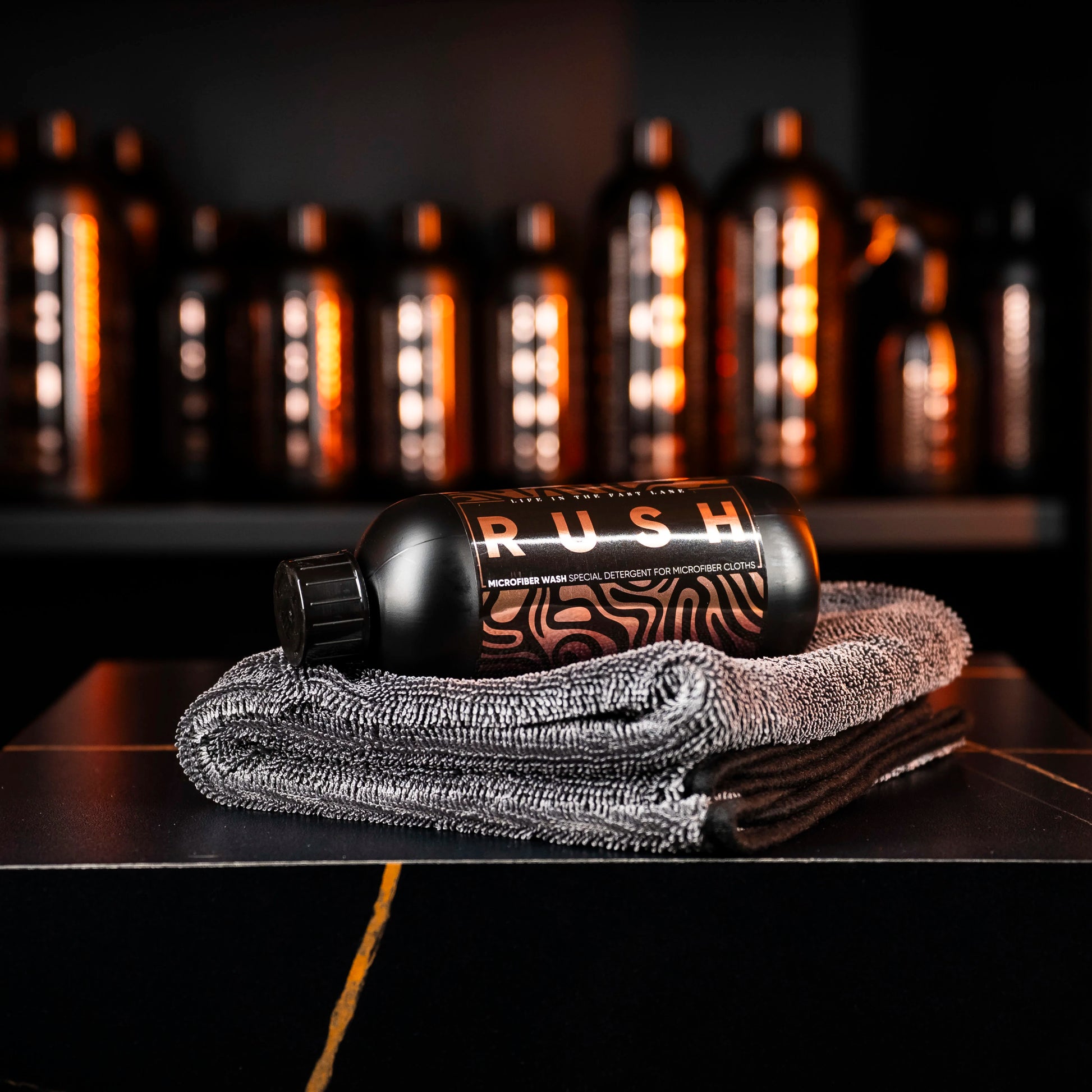 A black Perfect Drying Package bottle rests on folded gray microfiber towels, with more bottles blurred behind. The modern display and warm lighting emphasize the premium quality of this microfiber care product.