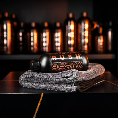 A black Perfect Drying Package bottle rests on folded gray microfiber towels, with more bottles blurred behind. The modern display and warm lighting emphasize the premium quality of this microfiber care product.