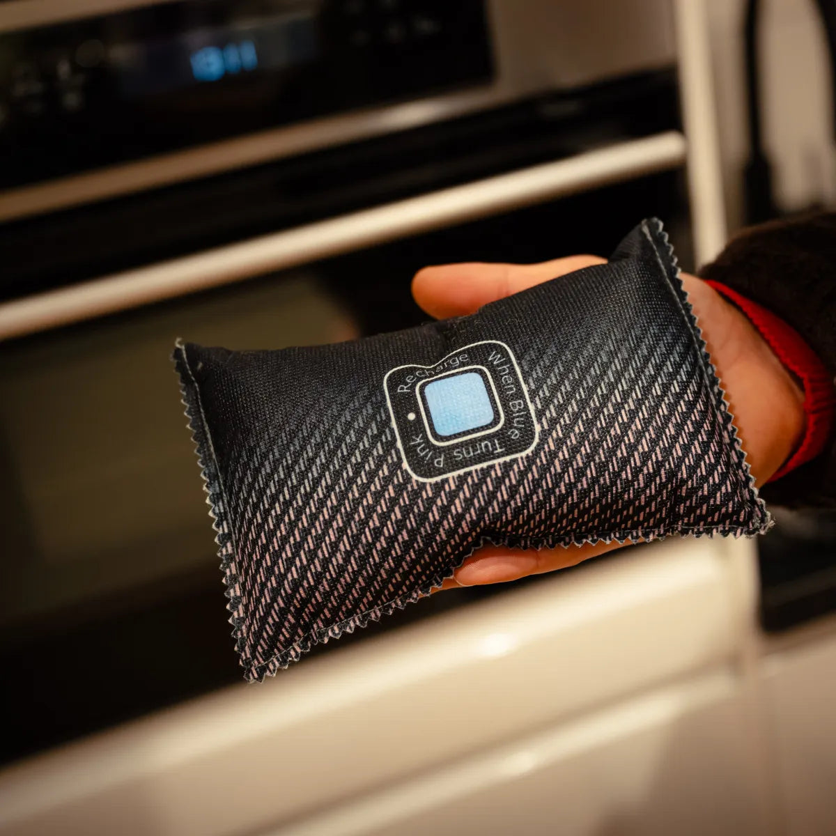 A hand holds the Auto Ontvochtiger 500gr, a rectangular black fabric pouch with blue square and patterned design, ideal for preventing fogged windows in your car with its effective moisture absorption.