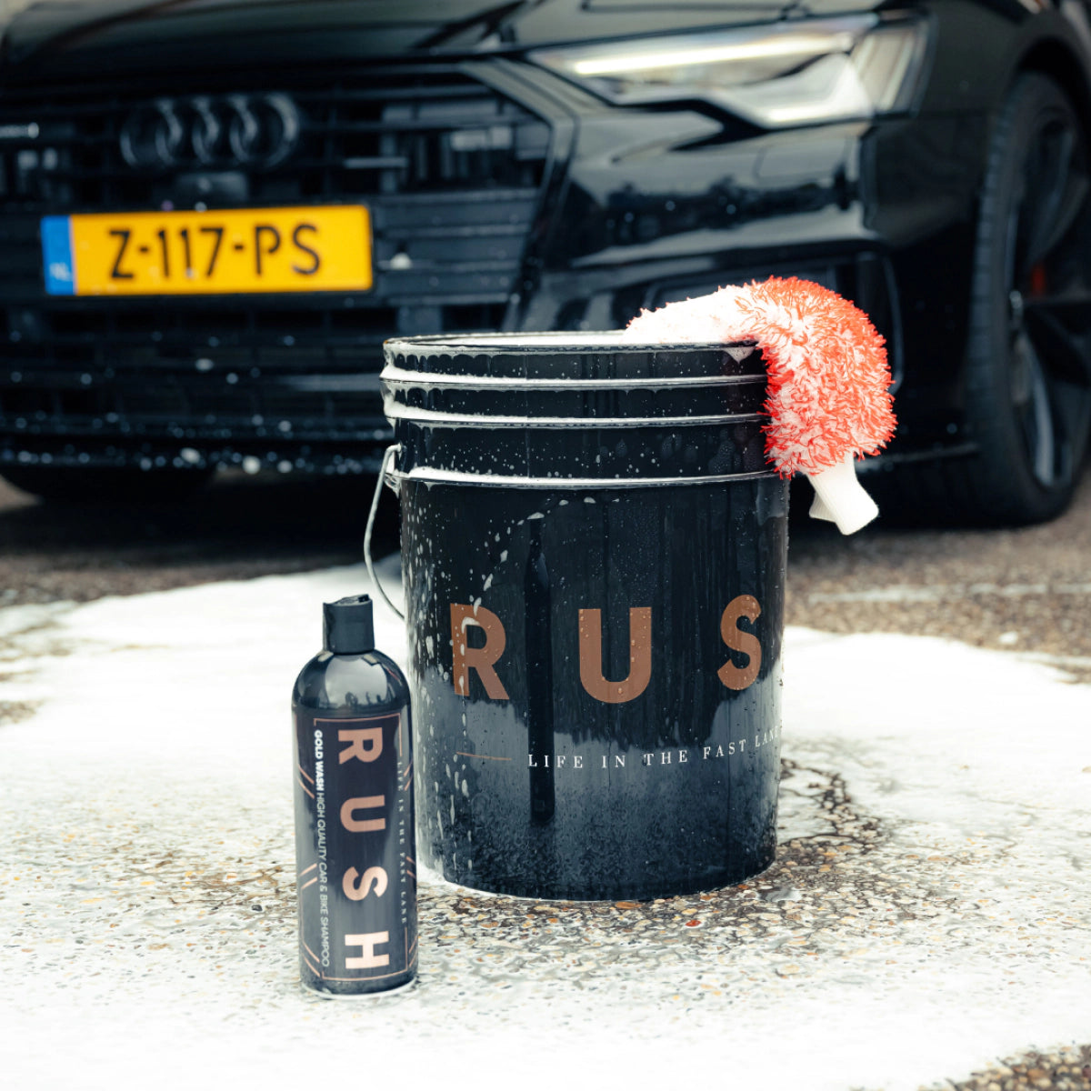 A black Detailing Bucket 20L with a red-and-white wash mitt and Grit Guard sits on wet pavement next to a RUSH car wash product. In the background, a black Audi with yellow plates highlights its use for the 2-emmer methode.