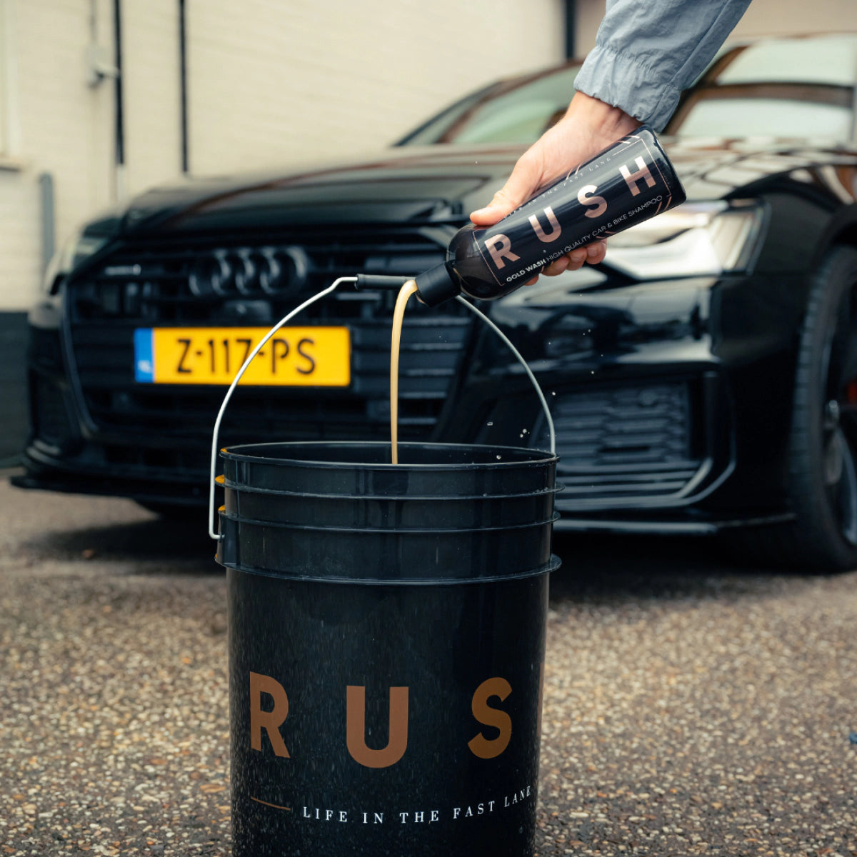 A person pours liquid from an Exterior Package bottle into a large black RUSH bucket, highlighting the premium Exterior Package for auto cleaning, with a sleek black car and yellow license plate in the background.