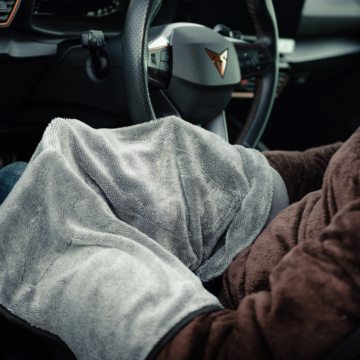A person sits in a car with a brown blanket on their lap and a grey microfiber cloth, holding the Window Clear Winter Kit near the steering wheel. The dashboard and steering wheel are visible.