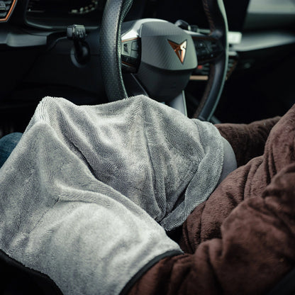 A person sits in a car with a brown blanket on their lap and a grey microfiber cloth, holding the Window Clear Winter Kit near the steering wheel. The dashboard and steering wheel are visible.