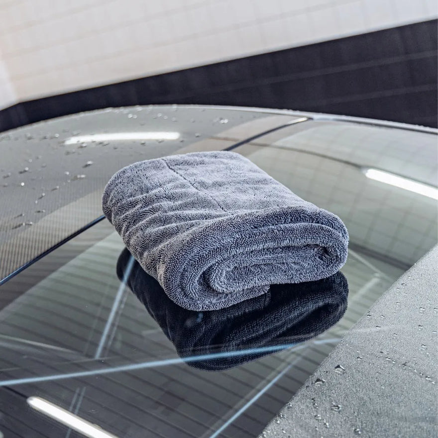A folded gray Master Drying Towel 1200 GSM rests on the wet rear windshield of a car, with water droplets and reflections of overhead lights visible on the glass.