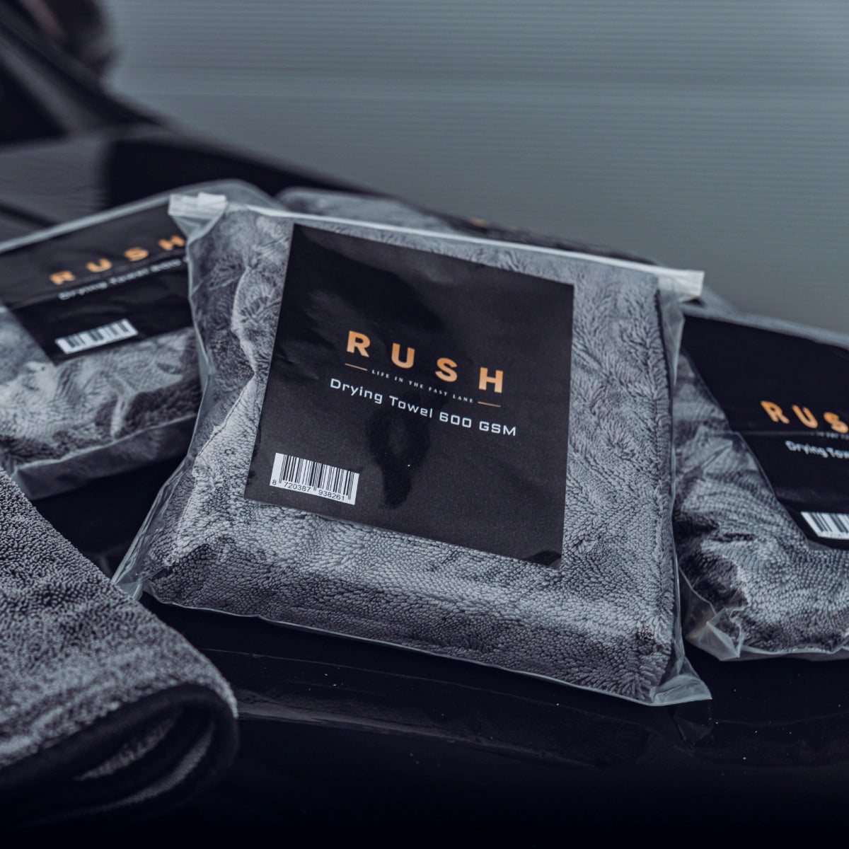 Three packaged Drying Towel 600 GSM towels are arranged on a dark, glossy surface, with part of an unpackaged gray towel visible in the corner.