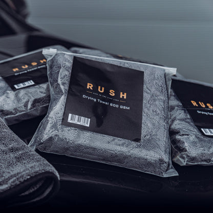 Three packaged Drying Towel 600 GSM towels are arranged on a dark, glossy surface, with part of an unpackaged gray towel visible in the corner.